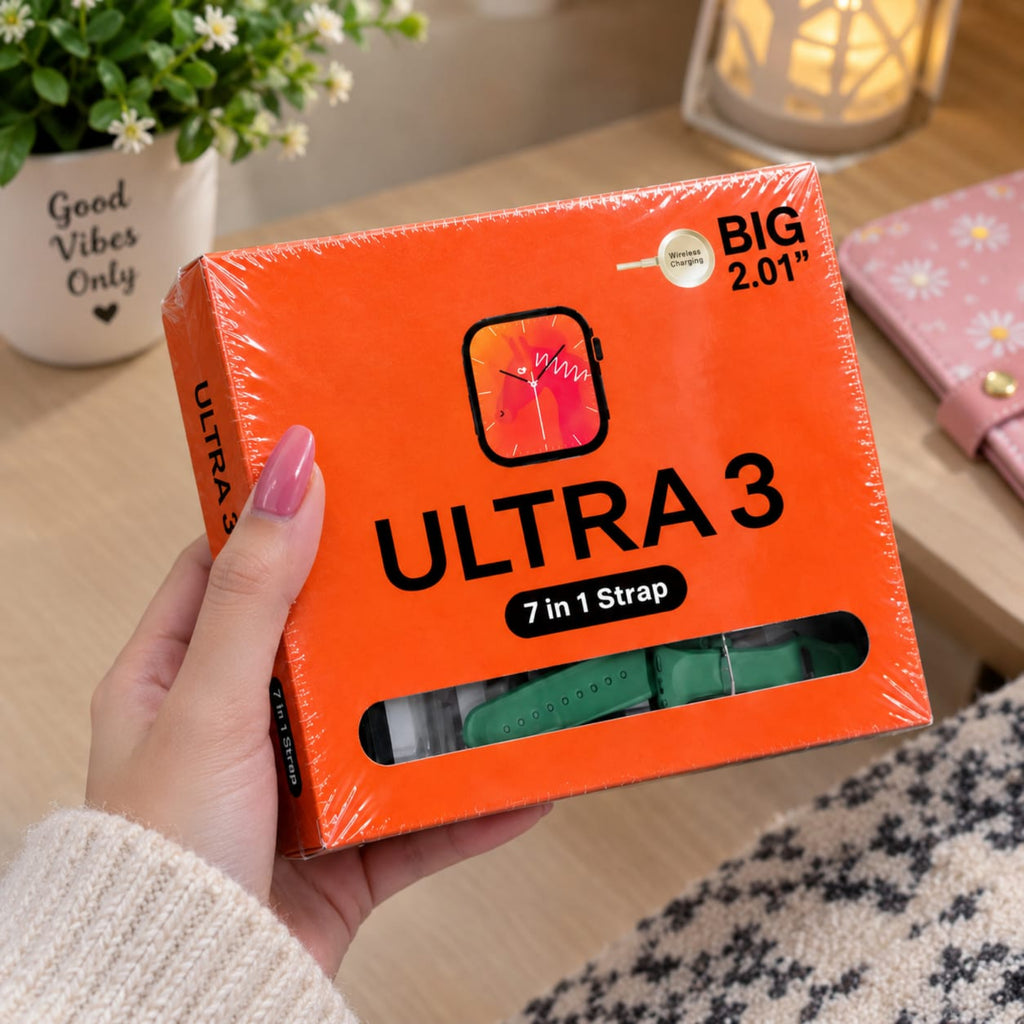 A hand with pink nail polish holds an orange box of ULTRA 3 7-in-1 watch strap in a cozy indoor setting with a wooden table, potted plant, and warm lamp in the background.