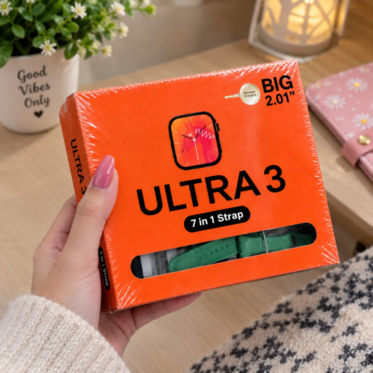 A hand with pink nail polish holds an orange box of ULTRA 3 7-in-1 watch strap in a cozy indoor setting with a wooden table, potted plant, and warm lamp in the background.