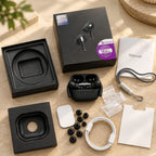 A pair of black wireless earbuds in an open charging case, displayed with accessories and packaging on a beige tabletop. The set includes a black retail box with earbud artwork, a manual, earbud cases, charging cable, ear tips, small plastic bags, and two black square inserts.