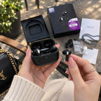 Black true wireless earbuds in an open charging case, held by two hands over a table with the product box, extra ear tips, a manual, and a black bag nearby.