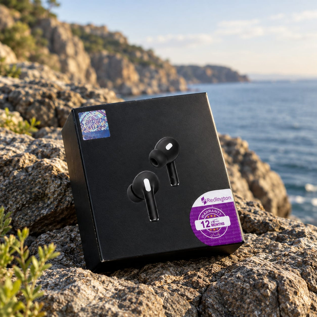 Black wireless earbud box with a pair of black and white earbuds printed on the front, placed on rocky coastal ground with the sea and cliffs blurred in the background. Purple Redington badges are visible on the box.