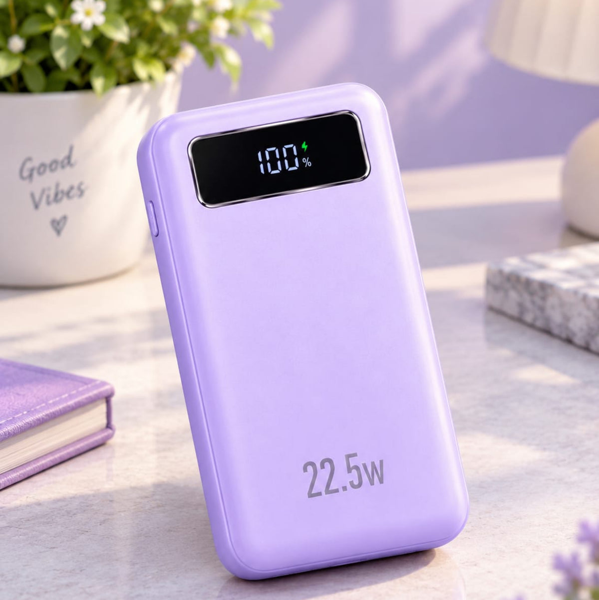 A lavender purple portable power bank with a digital display showing 100% battery level, standing on a light marble desk with a potted plant and notebook in the background.