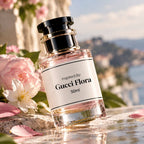 A 50ml bottle of Gucci Flora perfume, inspired by the fragrance, is displayed on a reflective stone surface next to pink and white flowers, with a scenic coastal background.