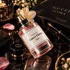 A 50ml bottle of Gucci Flora perfume lies on a glossy black marble surface, surrounded by luxury props including a Gucci Flora book, a black Gucci case, a pen, and white flowers. The product features a clear glass bottle filled with pink liquid, a white label with black text, and a black cap with gold detailing.